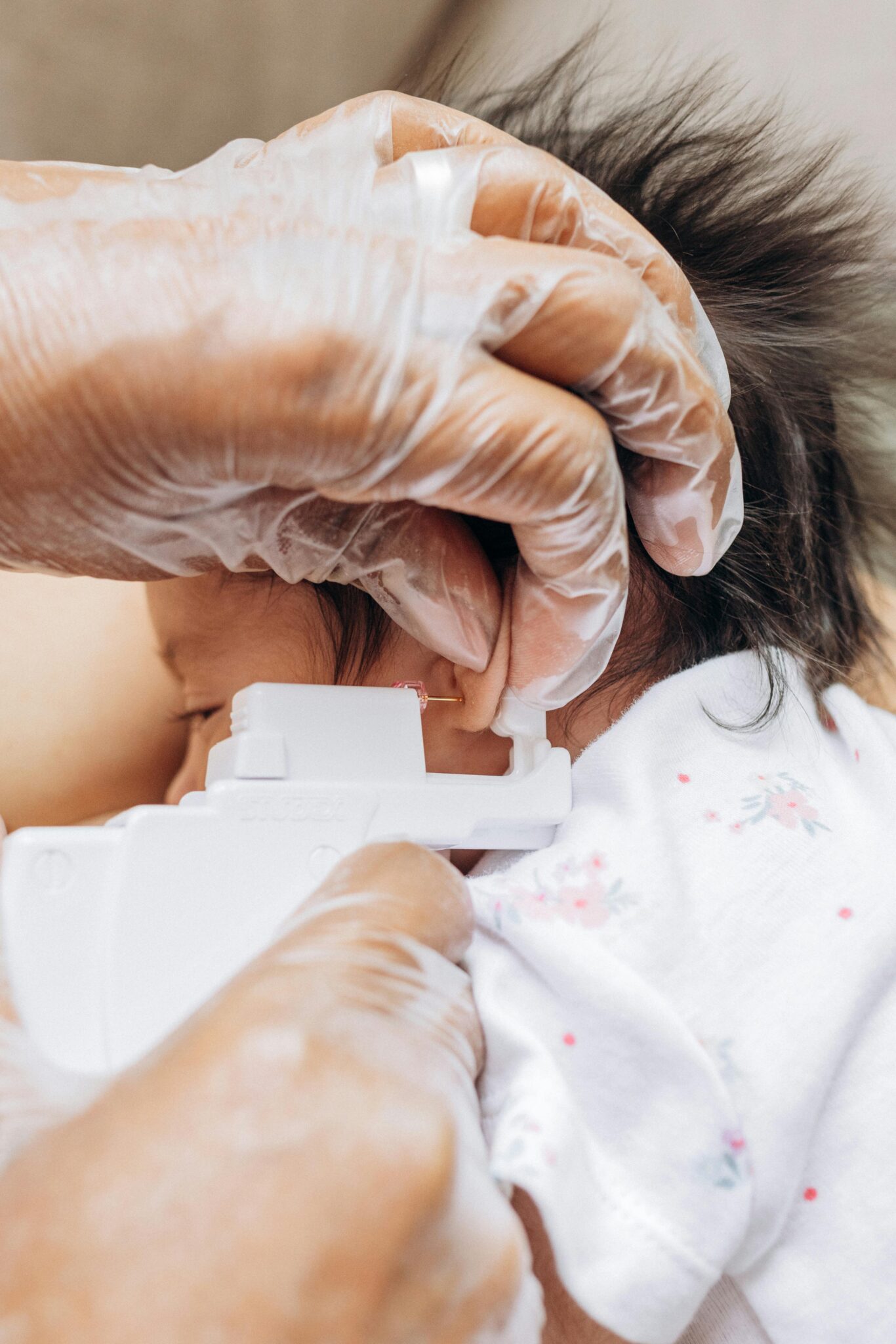 A close-up image of a baby's ear being pierced indoors, showing gloved hands and a piercing tool.